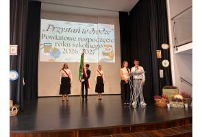 Poczet sztandarowy stoi na scenie podczas uroczystości rozpoczęcia roku szkolnego. Po prawej stronie dwoje uczniów znajduje się przy mównicy, a jeden z nich przemawia do mikrofonu.