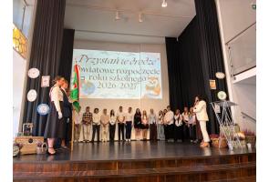 Grupa uczniów stoi w szeregu na scenie podczas uroczystości rozpoczęcia roku szkolnego. Po lewej stronie widoczny jest poczet sztandarowy z biało-czerwonymi szarfami, a po prawej kobieta przemawia do mikrofonu. W tle wyświetlana jest prezentacja, a po bokach sceny znajdują się zegary oraz dodatkowe dekoracje.