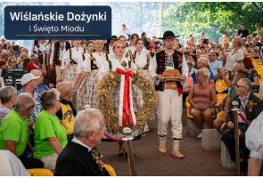 Grupa osób w tradycyjnych strojach regionalnych przechodzi pomiędzy rzędami siedzących uczestników wydarzenia. Na pierwszym planie kobieta niesie duży wieniec ozdobiony biało-czerwoną wstęgą, a obok mężczyzna trzyma bochenek chleba na drewnianej podstawie. W tle widoczna jest liczna publiczność oraz napis „Wiślańskie Dożynki i Święto Miodu”.