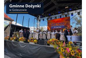 Grupa osób w tradycyjnych strojach regionalnych stoi na scenie. Przed sceną ustawiono liczne kompozycje z kolorowych kwiatów, a w tle widoczny jest ekran z napisem „Gminne Dożynki – Dni Gminy Goleszów 29–30.08.2026”.