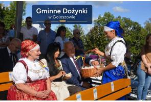 Kobieta w tradycyjnym stroju regionalnym stoi przy ławce i trzyma wiklinowy koszyk, z którego częstuje siedzące osoby. Obok widoczne są dwie kobiety, mężczyzna oraz zgromadzeni uczestnicy wydarzenia. W tle znajduje się napis „Gminne Dożynki w Goleszowie”.