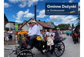 Osoby w tradycyjnych strojach regionalnych jadą udekorowaną bryczką zaprzężoną w konie. Pojazd ozdobiony jest kwiatami i kolorowymi wstążkami, a w tle widoczny jest korowód uczestników oraz zabudowania. W górnej części zdjęcia znajduje się napis „Gminne Dożynki w Goleszowie”.