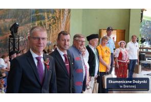 Grupa osób stoi w rzędzie. Część ma na sobie garnitury, a część tradycyjne stroje regionalne. Kilka osób trzyma papierowe torby. W tle widoczni są kolejni uczestnicy oraz dekoracja z krajobrazem i kłosami zbóż. W prawym dolnym rogu znajduje się napis „Ekumeniczne Dożynki w Brennej”.