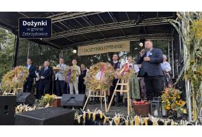 Grupa osób stoi na scenie wśród dożynkowych wieńców i dekoracji z kłosów zbóż, kukurydzy, kwiatów i owoców. Po prawej stronie mężczyzna przemawia do mikrofonu, a w tle widoczny jest napis „Dożynki Gminne”. W lewym górnym rogu znajduje się napis „Dożynki w gminie Zebrzydowice”.