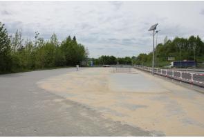 Widok na utwardzony plac przy drodze, częściowo pokryty piaskiem. Teren jest oddzielony barierkami, a obok znajdują się latarnie z panelami solarnymi. W tle widoczna jest jezdnia, zieleń oraz pojedyncze pojazdy.