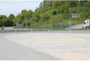 Widok na rozległy, utwardzony plac przy drodze, częściowo pokryty piaskiem. Wzdłuż krawędzi ustawiono metalowe stojaki oraz znaki drogowe. W tle widoczna jest jezdnia, samochód ciężarowy i porośnięte zielenią zbocze.