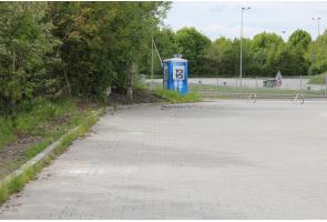 Widok na utwardzony plac otoczony zielenią. Po lewej stronie rosną drzewa i krzewy, a w tle znajduje się przenośna toaleta, ogrodzenie oraz elementy infrastruktury drogowej.