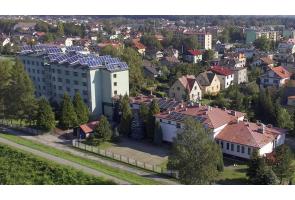 „Widok z lotu ptaka na kompleks budynków zlokalizowany w zabudowie miejskiej. Na pierwszym planie niższe budynki z czerwonymi dachami, w tle wysoki budynek z panelami fotowoltaicznymi na dachu.