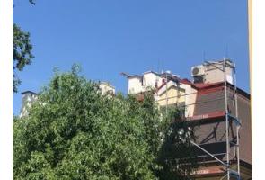 Widok na fragment budynku podczas prac remontowych. Przy elewacji ustawione jest rusztowanie, a na dachu widoczne są elementy konstrukcyjne i urządzenia techniczne. Dolną część budynku częściowo zasłaniają drzewa.