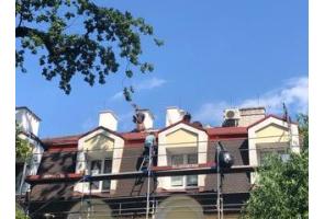 Widok na fragment budynku podczas prac remontowych na dachu. Przy elewacji ustawione jest rusztowanie, a na połaci dachowej pracują osoby wykonujące prace budowlane. W tle widoczne są kominy, okna dachowe oraz urządzenia techniczne.