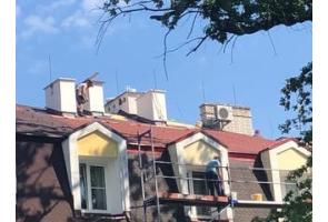 Widok na fragment budynku podczas prac remontowych dachu. Na połaci dachowej pracują osoby wykonujące prace budowlane, a przy elewacji ustawione jest rusztowanie. W tle widoczne są kominy, lukarny oraz urządzenie techniczne zamontowane na dachu.