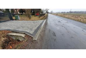 Droga asfaltowa z wjazdem z kostki brukowej prowadzącym do posesji po lewej stronie. Widoczny betonowy przepust pod zjazdem. Po prawej stronie pola uprawne. Nawierzchnia drogi wilgotna, niebo zachmurzone.