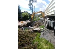 Prace ziemne przy drodze z użyciem koparki i samochodu ciężarowego. Widoczne elementy betonowe przygotowane do montażu. Po lewej stronie posesje, po prawej pas zieleni. Teren wykopu częściowo uprzątnięty, robotnicy w trakcie pracy.