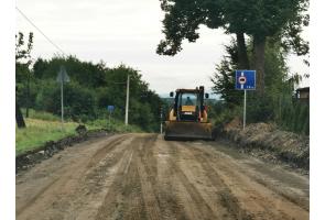Droga w trakcie remontu, nawierzchnia przygotowana do dalszych prac. Na środku jezdni spycharka. Po obu stronach drogi widoczne znaki drogowe oraz zieleń. Brak ruchu pojazdów, teren budowy.