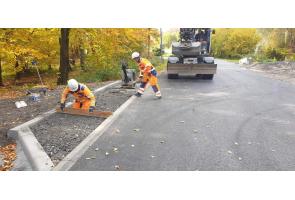 Prace wykończeniowe przy nowo wybudowanej drodze. Dwóch pracowników w odzieży ochronnej montuje elementy infrastruktury drogowej przy krawężniku. Widoczna świeżo ułożona nawierzchnia asfaltowa oraz sprzęt budowlany w tle. Otoczenie zalesione, liście w kolorach jesieni.