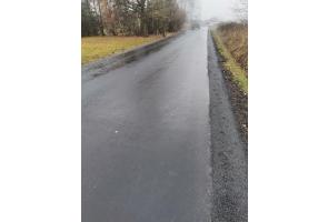 Droga asfaltowa po zakończonym remoncie. Nawierzchnia równa i ciemna, świeżo ułożona. Po obu stronach drogi pobocza gruntowe z roślinnością. W tle widoczność ograniczona przez mgłę. Otoczenie wiejskie z pojedynczymi drzewami.