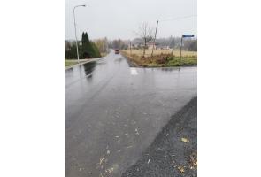 Skrzyżowanie dróg asfaltowych po zakończeniu prac remontowych. Nawierzchnia równa i wilgotna. Po lewej stronie lampa uliczna i posesja, po prawej pole uprawne oraz znak drogowy z nazwą ulicy. Teren otwarty, widoczność dobra mimo pochmurnej pogody.