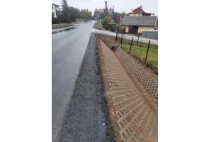 Droga asfaltowa po remoncie z utwardzonym poboczem. Po prawej stronie wykonany rów odwadniający z betonowej kratki typu ażurowego oraz przepust pod zjazdem. W tle zabudowa jednorodzinna i znaki drogowe. Nawierzchnia wilgotna, pogoda pochmurna.