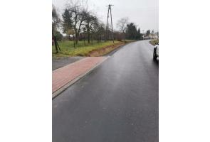 Droga asfaltowa po remoncie z przylegającym chodnikiem z kostki brukowej po lewej stronie. Po prawej stronie fragment pojazdu, dalej teren zielony i drzewa. W oddali linie energetyczne i zabudowa jednorodzinna. Nawierzchnia wilgotna, pogoda pochmurna.