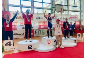 Ceremonia dekoracji podczas zawodów Olimpiad Specjalnych w hali sportowej. Szymon Machej stoi na najwyższym stopniu podium z medalami, obok pozostali zawodnicy i osoby towarzyszące.