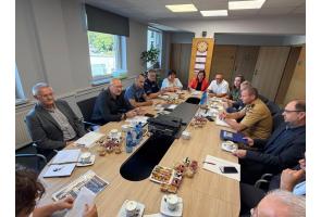 Uczestnicy comiesięcznej narady dotyczącej bezpieczeństwa siedzą przy stole konferencyjnym. W spotkaniu biorą udział przedstawiciele samorządu oraz różnych służb działających na terenie Powiatu Cieszyńskiego. Na stole znajdują się materiały robocze, notatniki i dokumenty.