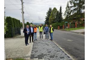 Sześciu mężczyzn stoi na chodniku przy wyremontowanej drodze. Dwóch z nich ma na sobie odblaskowe kamizelki, pozostali ubrani są w garnitury lub codzienne stroje. W tle widoczne są domy, żywopłoty, drzewa oraz słupy z przewodami.