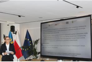 Kobieta stoi obok dużego ekranu i trzyma w dłoniach kartki. Na ekranie wyświetlana jest prezentacja dotycząca projektu związanego z odnawialnymi źródłami energii, poprawą jakości powietrza i budową instalacji fotowoltaicznych. W tle widoczne są flagi.