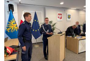 Umundurowany komendant przemawia przy mównicy podczas sesji rady powiatu; obok stoi drugi policjant, a na stole leży bukiet kwiatów.