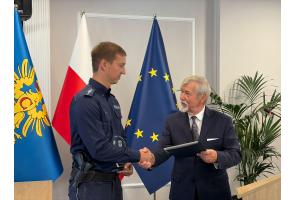 Aspirant Rafał Górczyński odbiera wyróżnienie i ściska dłoń Stanisława Kubiciusa; w tle flagi Polski, Unii Europejskiej i powiatu.