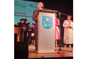 Przewodniczący Rady Powiatu przemawia przy mównicy podczas inauguracji roku akademickiego; za nim stoją słuchacze Uniwersytetu Trzeciego Wieku w biretach.