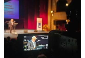 Kamera rejestruje starszego mężczyznę przemawiającego przy mównicy podczas inauguracji Uniwersytetu Trzeciego Wieku.