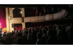 Słuchacze podczas inauguracji Uniwersytetu Trzeciego Wieku w ozdobnej sali teatralnej; publiczność siedzi na widowni, a przy scenie stoi bukiet kwiatów.
