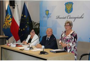 Podczas VIII sesji Rady Seniorów kobieta przemawia przy stole obrad; obok siedzą dwoje uczestników, w tle flagi Polski, UE i Powiatu Cieszyńskiego.