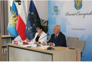 Dwoje uczestników VIII sesji Rady Seniorów Powiatu Cieszyńskiego przy stole obrad; przed nimi mikrofony i dokumenty, za nimi flagi oraz herb powiatu.