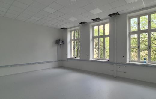 Wyremontowane pomieszczenie z jasnymi ścianami, dużymi oknami i nową podłogą. Przy ścianach widoczne są elementy instalacji, a w suficie znajdują się częściowo otwarte panele.