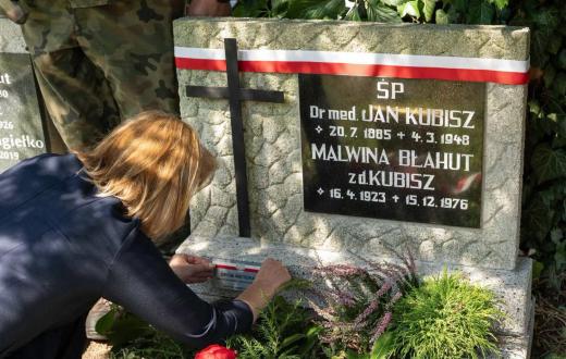 Kobieta przy grobie ozdobionym biało-czerwoną wstęgą umieszcza oznaczenie na płycie nagrobnej. Wokół widoczne są rośliny oraz kwiaty.