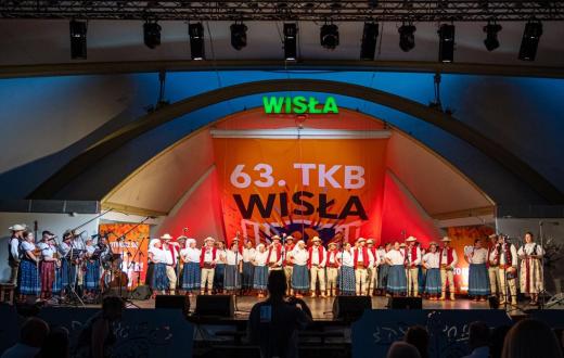 Grupa osób w strojach regionalnych stoi w rzędzie na scenie. Po lewej stronie widoczni są muzycy z instrumentami, a po prawej dwie kobiety stoją przy mikrofonach. W tle znajduje się napis „63. TKB Wisła”. Oświetlenie sali jest ciemne, a światło sceniczne pada głównie na stojące osoby.