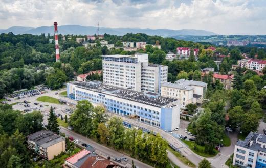 Rozległy kompleks budynków otoczony zielenią, widziany z lotu ptaka. Wokół znajdują się drogi, parkingi i zabudowa miejska, a w tle widoczne są wzgórza i góry.