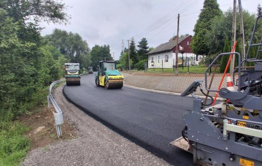 Maszyny drogowe układają i zagęszczają nową warstwę asfaltu na jezdni. Wzdłuż drogi widoczne są bariery ochronne, chodnik oraz zabudowania otoczone zielenią.