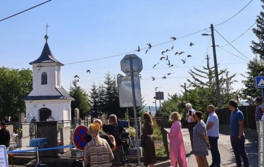 Grupa osób stoi przy ogrodzeniu w pobliżu niewielkiej kaplicy. Nad drogą przelatuje stado ptaków, a obok ustawiony jest sprzęt filmowy i znaki drogowe.
