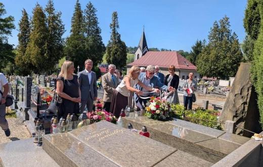 Grupa osób stoi przy grobie na cmentarzu. Dwie osoby na pierwszym planie układają kwiaty i znicze, a pozostali stoją obok. W tle widoczne są nagrobki, drzewa i budynek z wieżyczką.