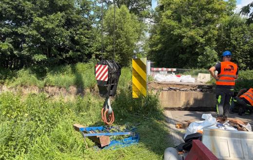 Prace przy uszkodzonym moście. Widoczny jest hak dźwigu, bariery zabezpieczające, materiały budowlane oraz pracownik w odzieży odblaskowej. Wokół znajduje się gęsta zieleń.