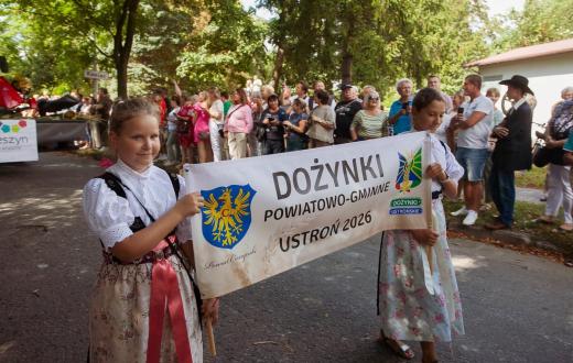 Dwie dziewczynki w strojach regionalnych niosą baner z napisem „Dożynki Powiatowo-Gminne Ustroń 2026”. W tle stoją licznie zgromadzeni uczestnicy i widzowie korowodu.