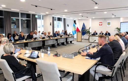 Widok na salę sesyjną podczas obrad. Uczestnicy siedzą przy ustawionych w podkowę stołach, przed nimi znajdują się mikrofony, tablety i tabliczki z nazwiskami. W centralnej części sali widoczne są mównica oraz flagi, a na ścianie godło Polski i herb Powiatu Cieszyńskiego.