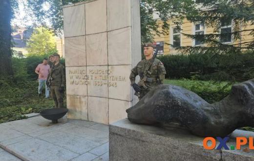 Pomnik z jasną kamienną płytą z napisem oraz ciemną rzeźbą umieszczoną na cokole. Po obu stronach monumentu stoją żołnierze w mundurach, a w tle widoczne są drzewa, krzewy i budynek.