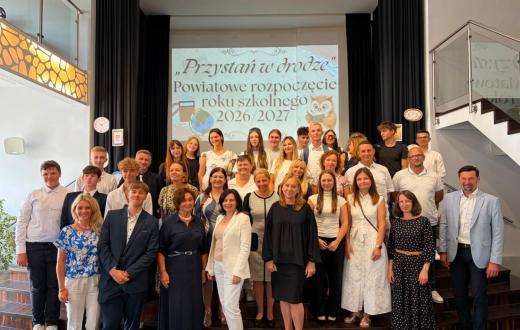 Grupa uczniów i dorosłych pozuje do wspólnego zdjęcia na schodach przed sceną. W tle wyświetlana jest prezentacja dotycząca powiatowego rozpoczęcia roku szkolnego 2026/2027. Po bokach sceny widoczne są zegary oraz kartki z cyframi.