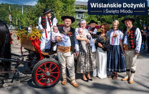 Grupa dorosłych i dzieci pozuje do wspólnego zdjęcia obok udekorowanego wozu zaprzęgowego. Część osób ma na sobie tradycyjne stroje regionalne, a niektórzy trzymają dzieci na rękach. W tle widoczne są drzewa, zabudowania oraz napis „Wiślańskie Dożynki i Święto Miodu”.