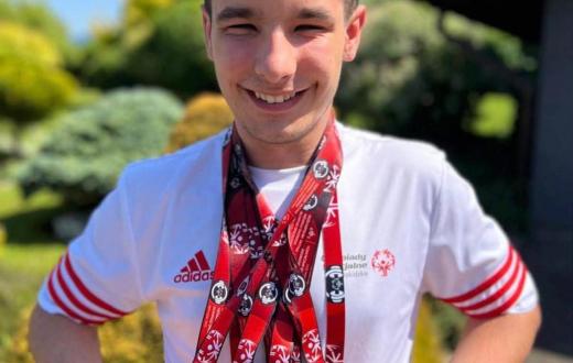 Szymon Machej stoi na zewnątrz w koszulce Olimpiad Specjalnych, uśmiecha się i prezentuje zawieszone na szyi medale zdobyte podczas zawodów.