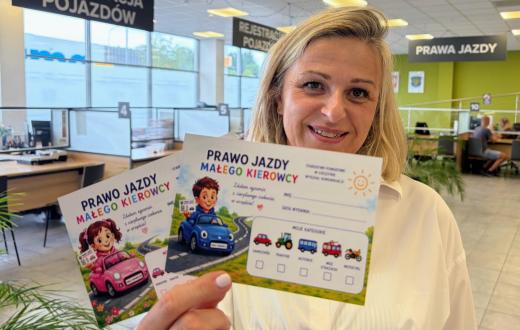 Kobieta trzyma w dłoni materiały zatytułowane „Prawo jazdy małego kierowcy”, ozdobione ilustracjami dzieci i kolorowych samochodów. W tle widoczne są stanowiska obsługi, duże okna oraz tablice z napisami „Rejestracja pojazdów” i „Prawa jazdy”.