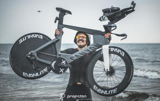 Mężczyzna w stroju sportowym i kasku unosi nad głową rower, stojąc w płytkiej wodzie przy brzegu morza. Ma na sobie sportowe okulary, a na twarzy widoczny jest uśmiech. W tle rozciąga się morze i pochmurne niebo.